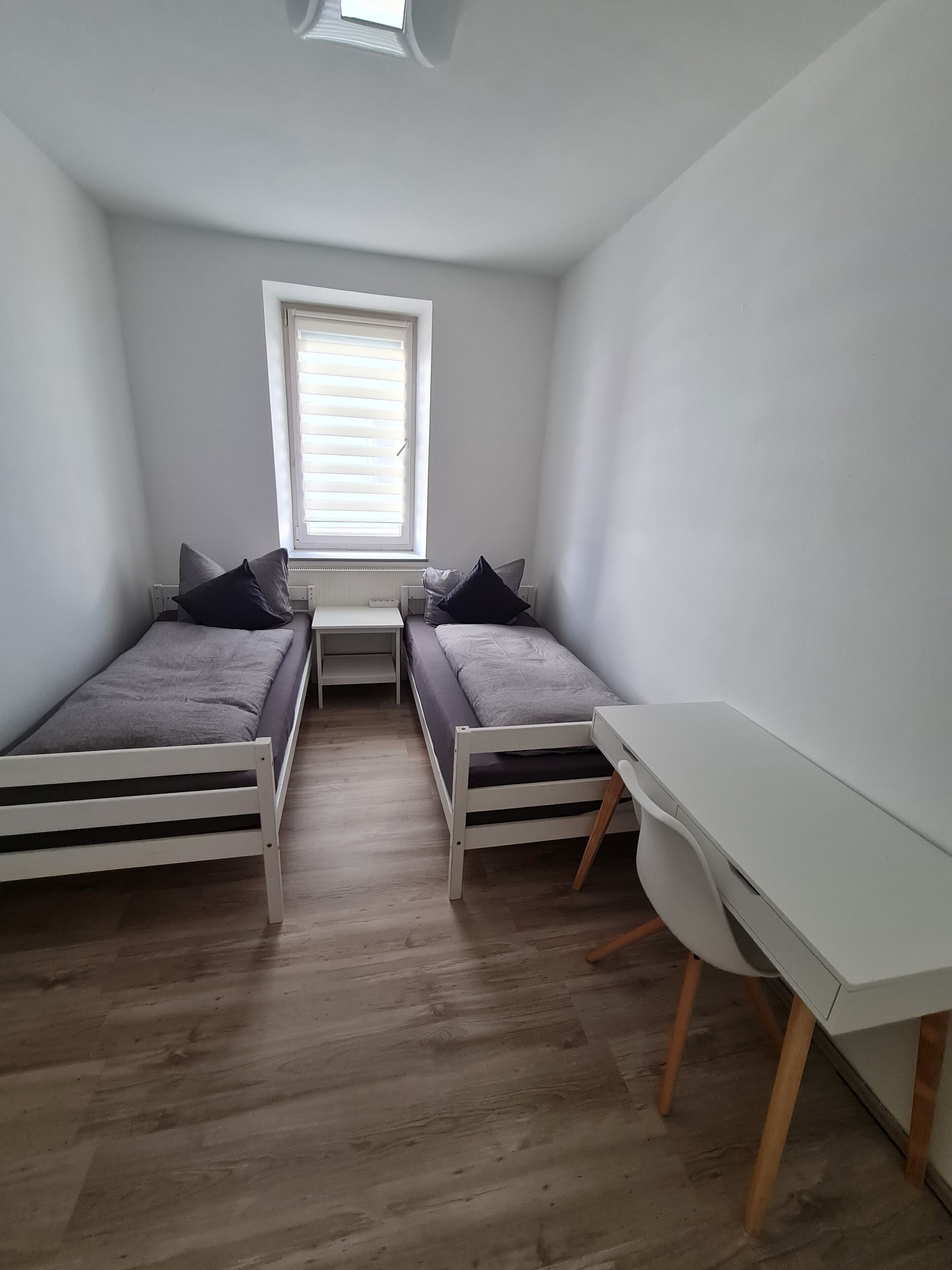 A small, modern room with two single beds, a white desk, and a window with blinds.