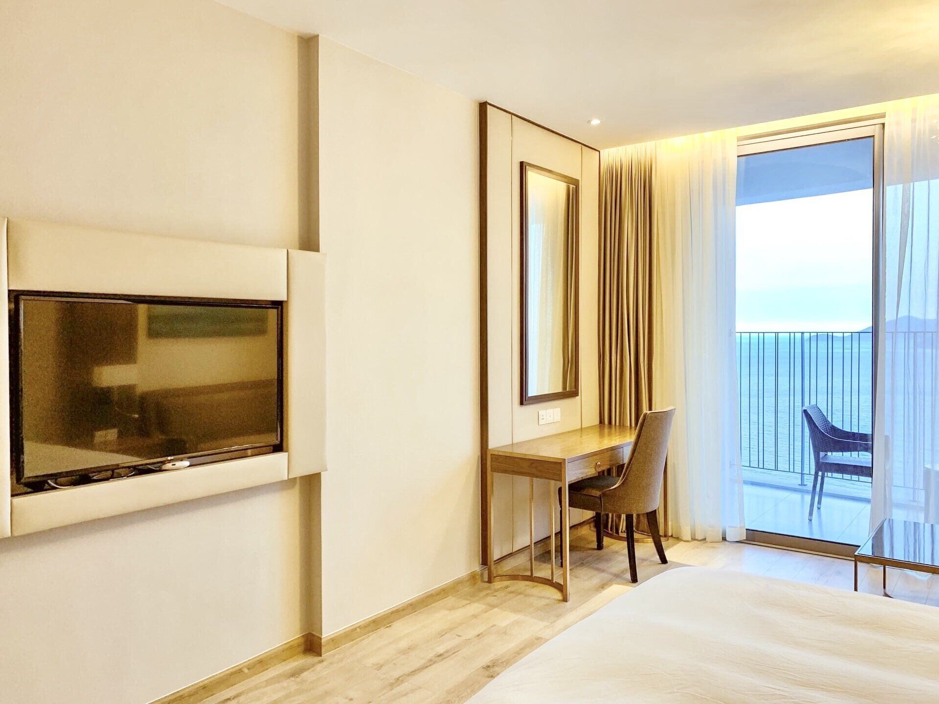 A hotel room with a bed, a desk, a chair, a TV, and a balcony with a view of the sea.