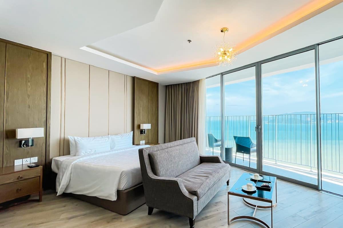A modern hotel room with a large bed, a sofa, a small table, and a balcony with a view of the sea.