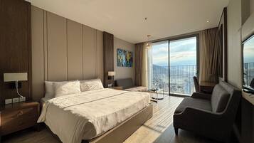 A modern hotel room with a large bed, a seating area, and a city view through large windows.