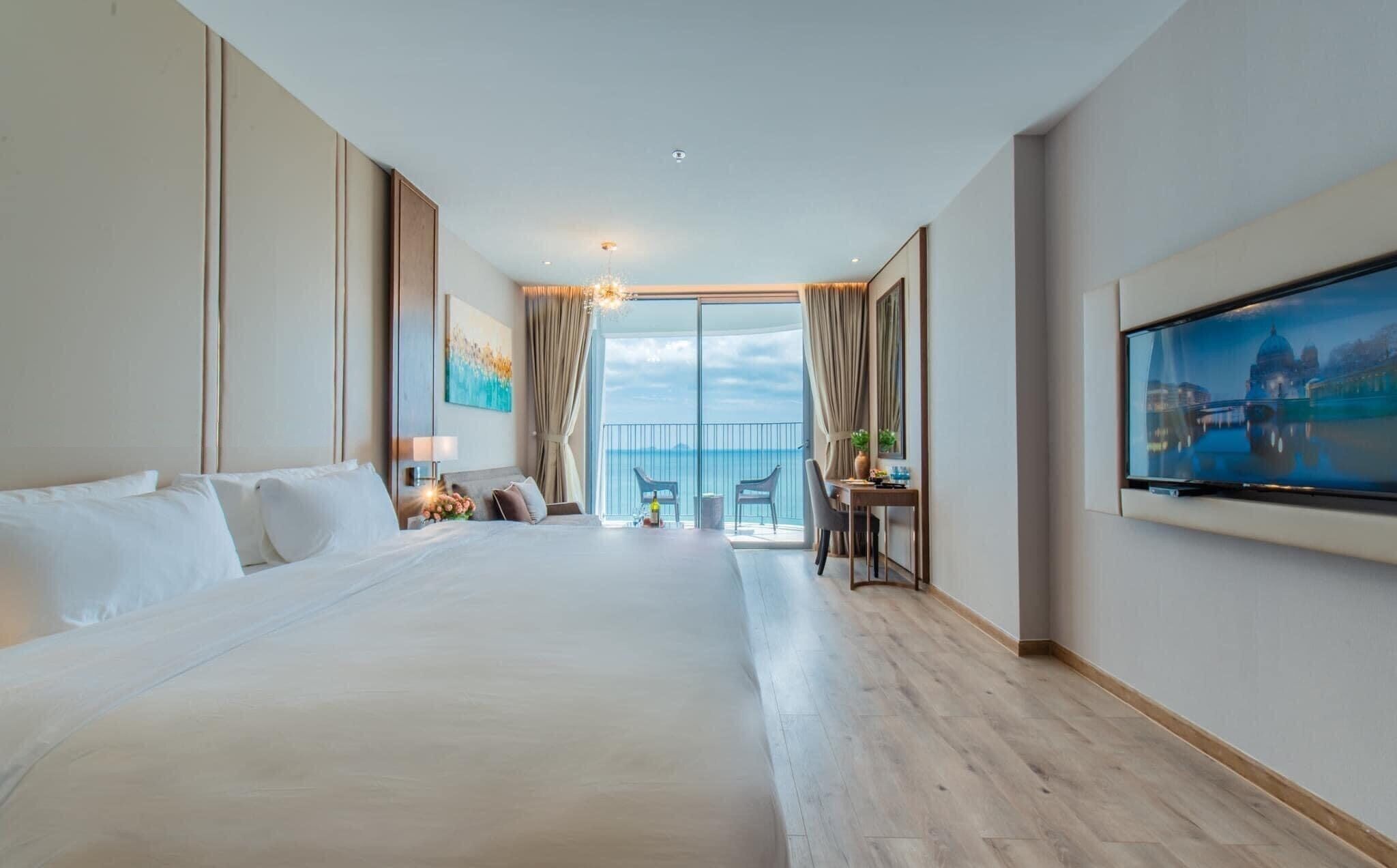 A hotel room with a large bed, a desk with a chair, a TV, and a balcony with a view of the sea.