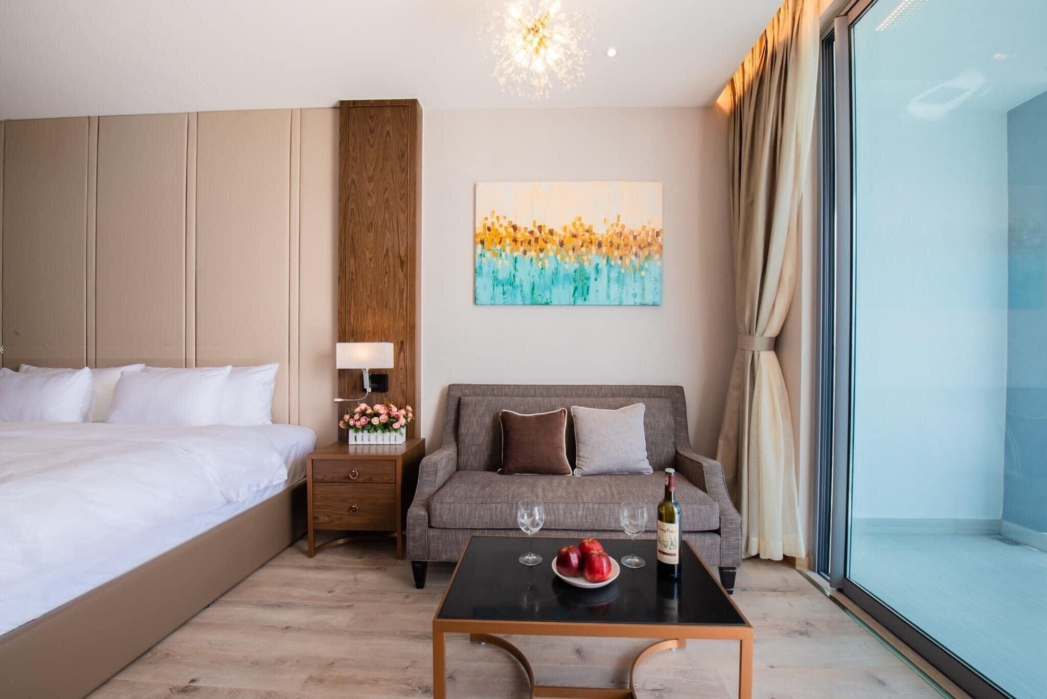 A hotel room with a bed, a sofa, a small table with a bottle and glasses, a nightstand with a lamp and flowers, and a painting on the wall.