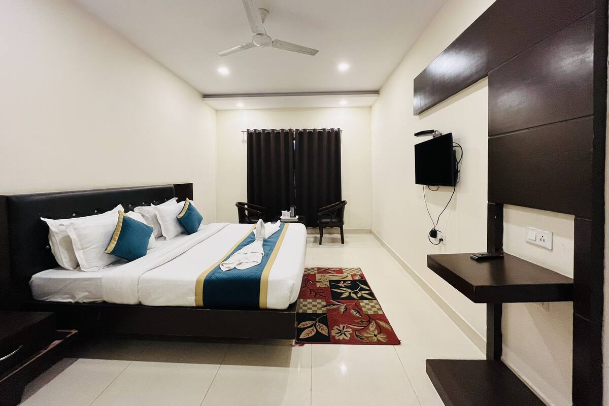 Quarto de hotel com cama, escrivaninha, cadeira, ventoinha no teto, televisão e janela com cortinas.