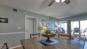 A dining area with a wooden table, chairs, and a bowl of fruit. There is a door leading to another room, a window with a view, and a ceiling fan. - Sea Banks 204, Two Bedroom, Two Bathroom, Oceanfront (Garden City Beach)