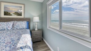 A bedroom with a bed, a nightstand, a lamp, a framed picture, and a large window with a beach view. - Sea Banks 204, Two Bedroom, Two Bathroom, Oceanfront (Garden City Beach)