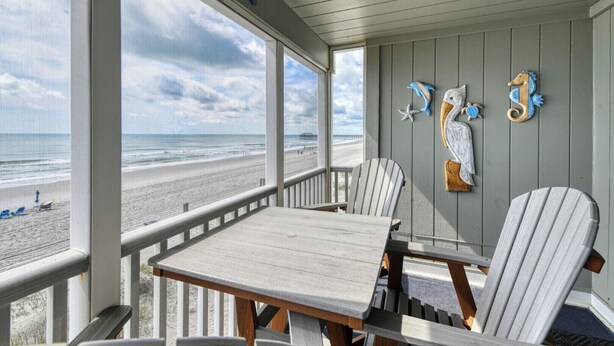 A covered outdoor seating area with a view of the beach, featuring a table and chairs, and decorative wall art. - Sea Banks 204, Two Bedroom, Two Bathroom, Oceanfront (Garden City Beach)