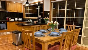 Un comedor con una mesa de madera dispuesta para seis personas, rodeada de sillas de madera con asientos acolchados. Se observa un cesto de basura a la izquierda de la mesa.