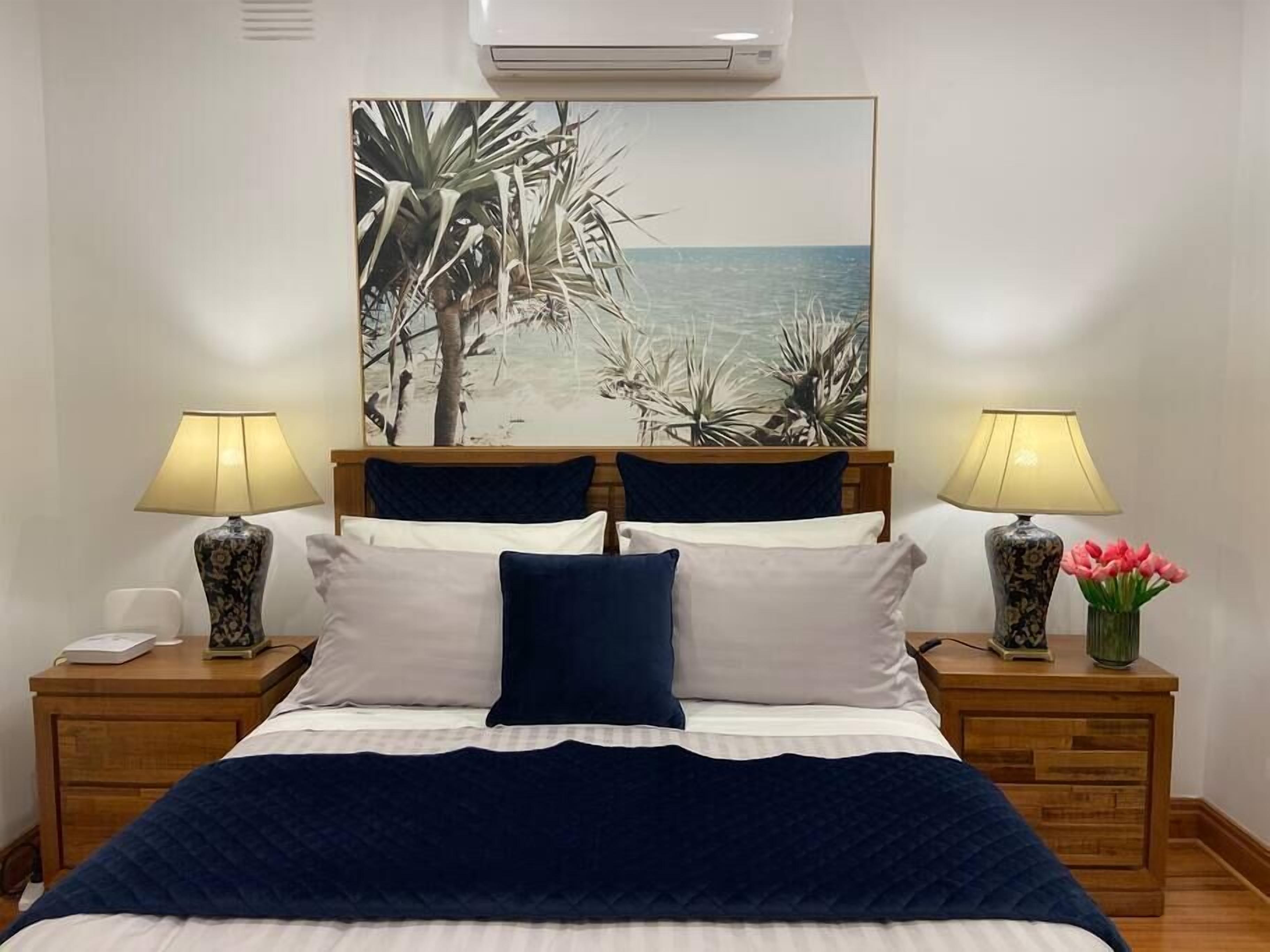 A neatly made bed with a blue blanket, two bedside lamps, and a painting of a beach scene above it.
