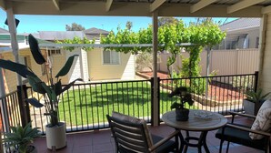 Un porche cubierto con una mesa y sillas, rodeado de plantas en macetas y vistas a un jardín con césped y una valla.