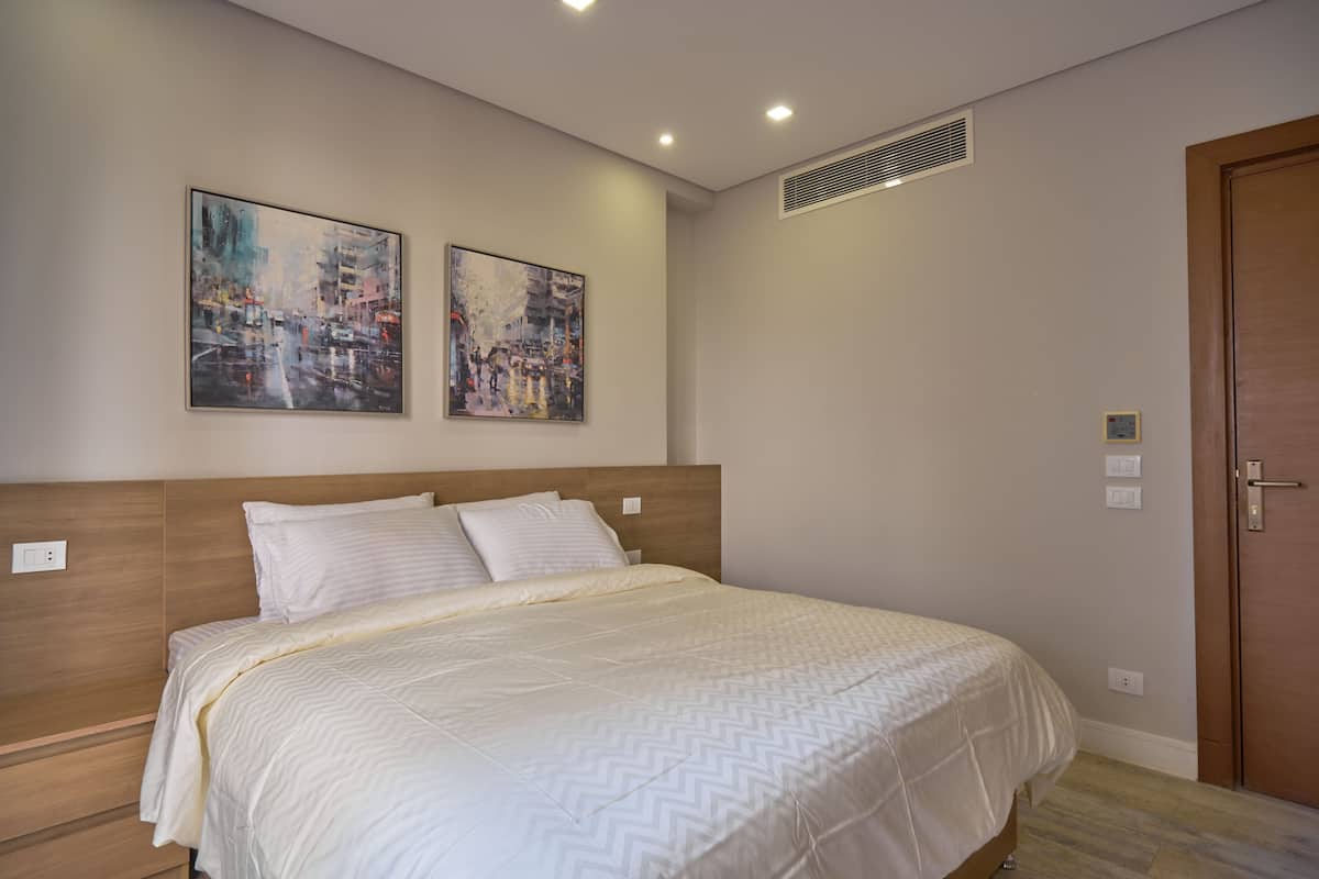 A bedroom with a large bed, two wall-mounted paintings, a wooden headboard, a door, and a vent.