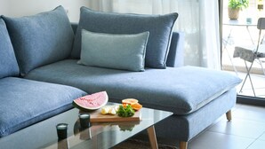 Una sala de estar moderna con un sofá gris, una mesa de centro de vidrio y una pequeña mesa de comedor con sandía, naranjas y una botella.