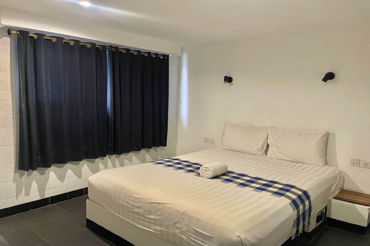 Una habitación de hotel con cama, mesita de noche, ventana con cortinas y lámpara de pared.