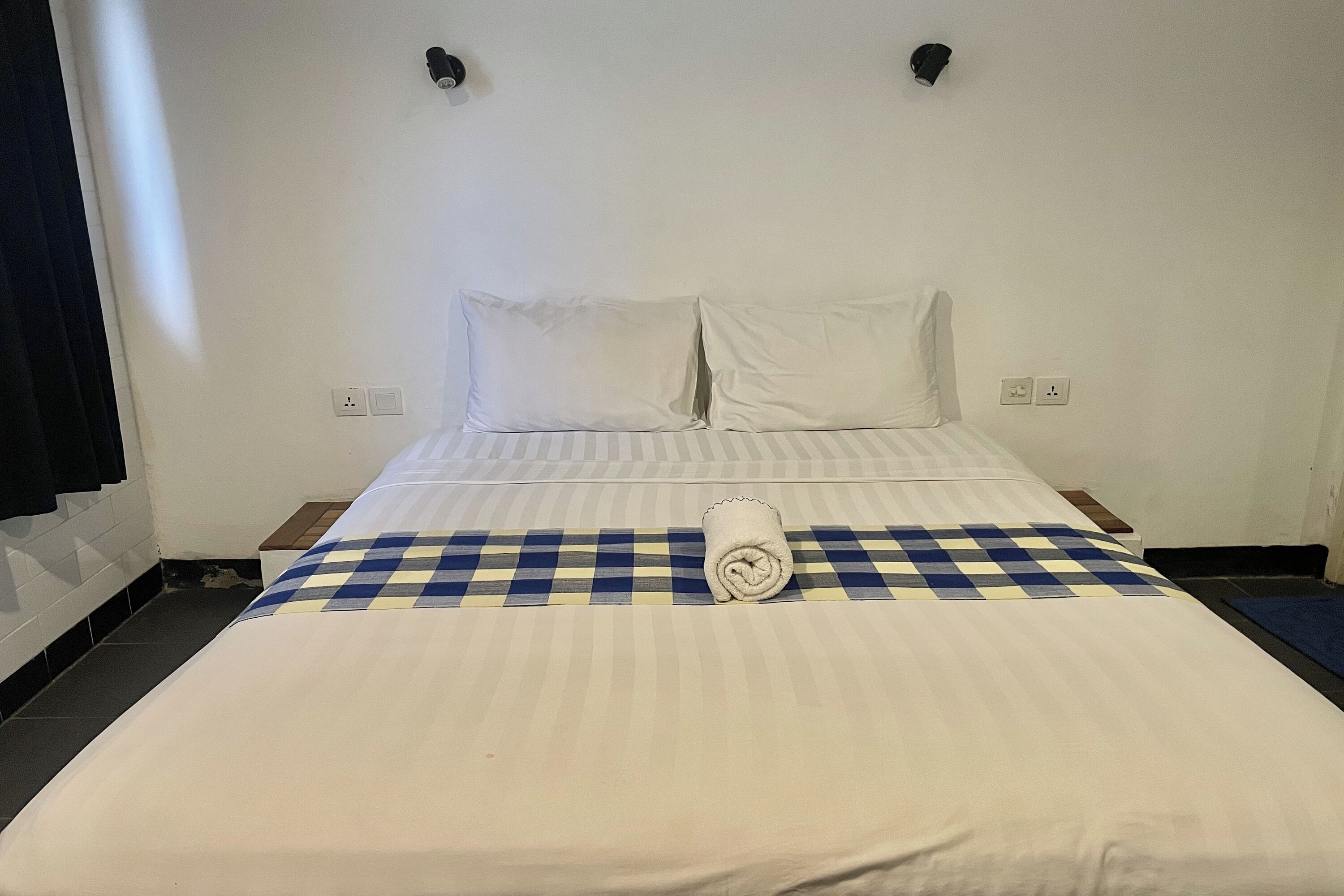 A hotel room with a neatly made bed, a checkered blanket, and a rolled towel on the bed.
