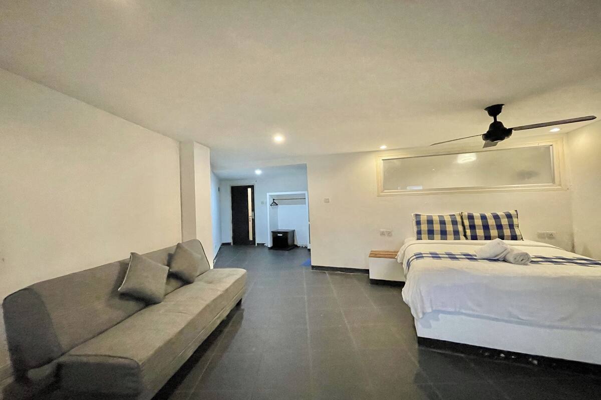 Habitación de hotel moderna con un sofá gris, una cama con colcha azul y blanca a cuadros y un ventilador de techo.