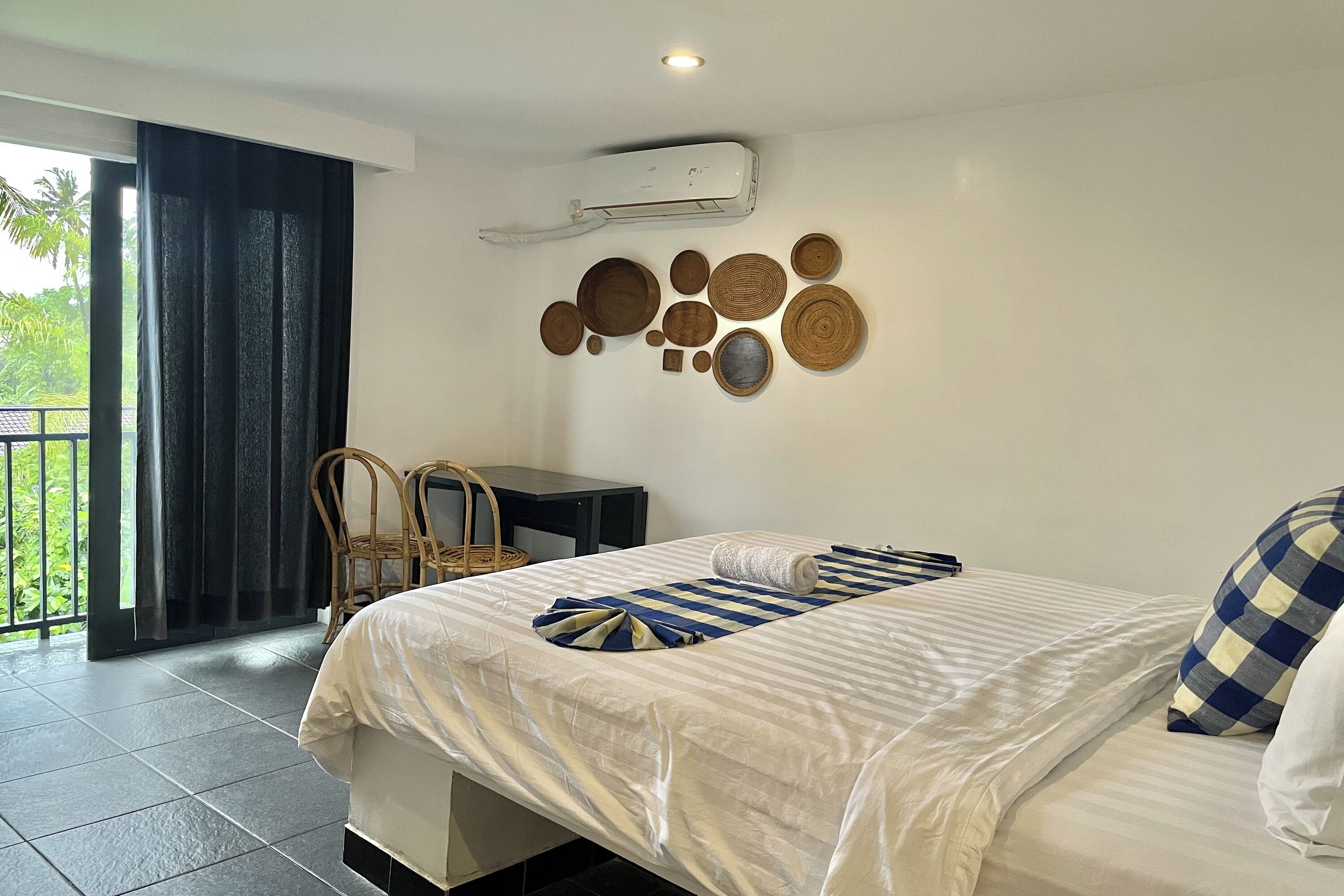 A hotel room with a bed, a wall-mounted air conditioner, a small table, chairs, and a balcony with a view of greenery.