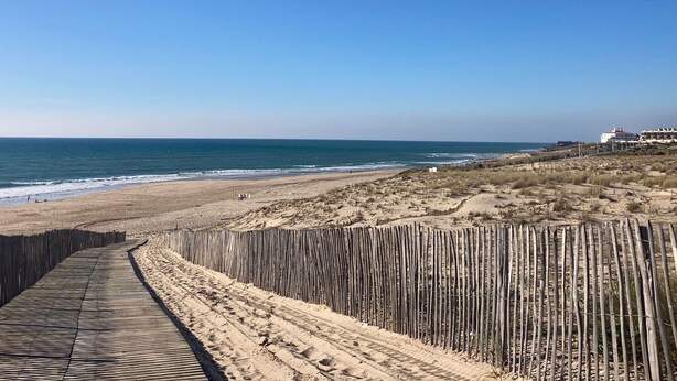 Een strand met een houten vogelbeschermingsgang die naar de kust leidt, zandstrand en gebouwen op de achtergrond.