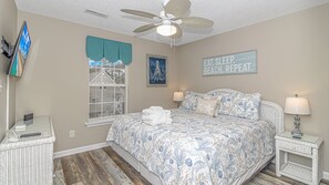 Une chambre à coucher comprenant un lit, deux tables de chevet, un ventilateur de plafond, une fenêtre avec stores, un téléviseur fixé au mur et une citation inspirante sur le mur.