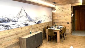Un intérieur en bois avec une table à manger dressée pour quatre personnes, un tableau représentant une montagne accroché au mur et une porte en bois.