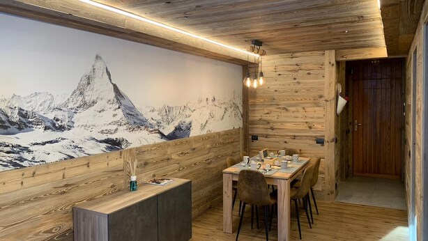 Un espace repas avec une table et des chaises en bois, une fresque représentant une montagne et un plafond en bois agrémenté de luminaires suspendus.