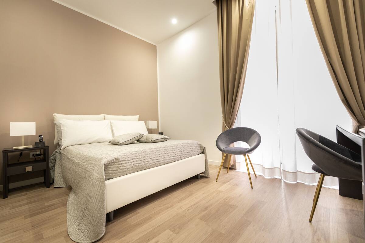 A modern bedroom with a bed, bedside tables, a chair, and a large window with curtains.