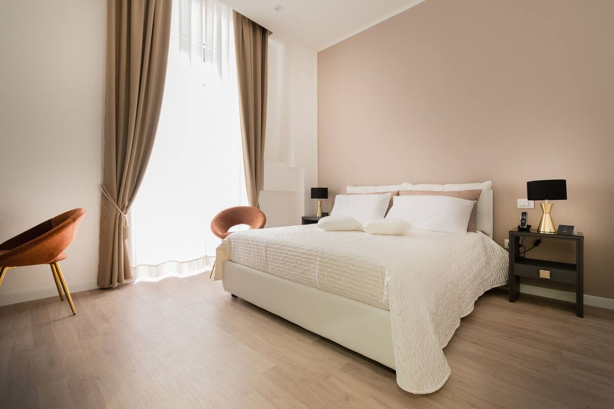 A modern bedroom with a large bed, two bedside tables with lamps, a chair, and a window with curtains.
