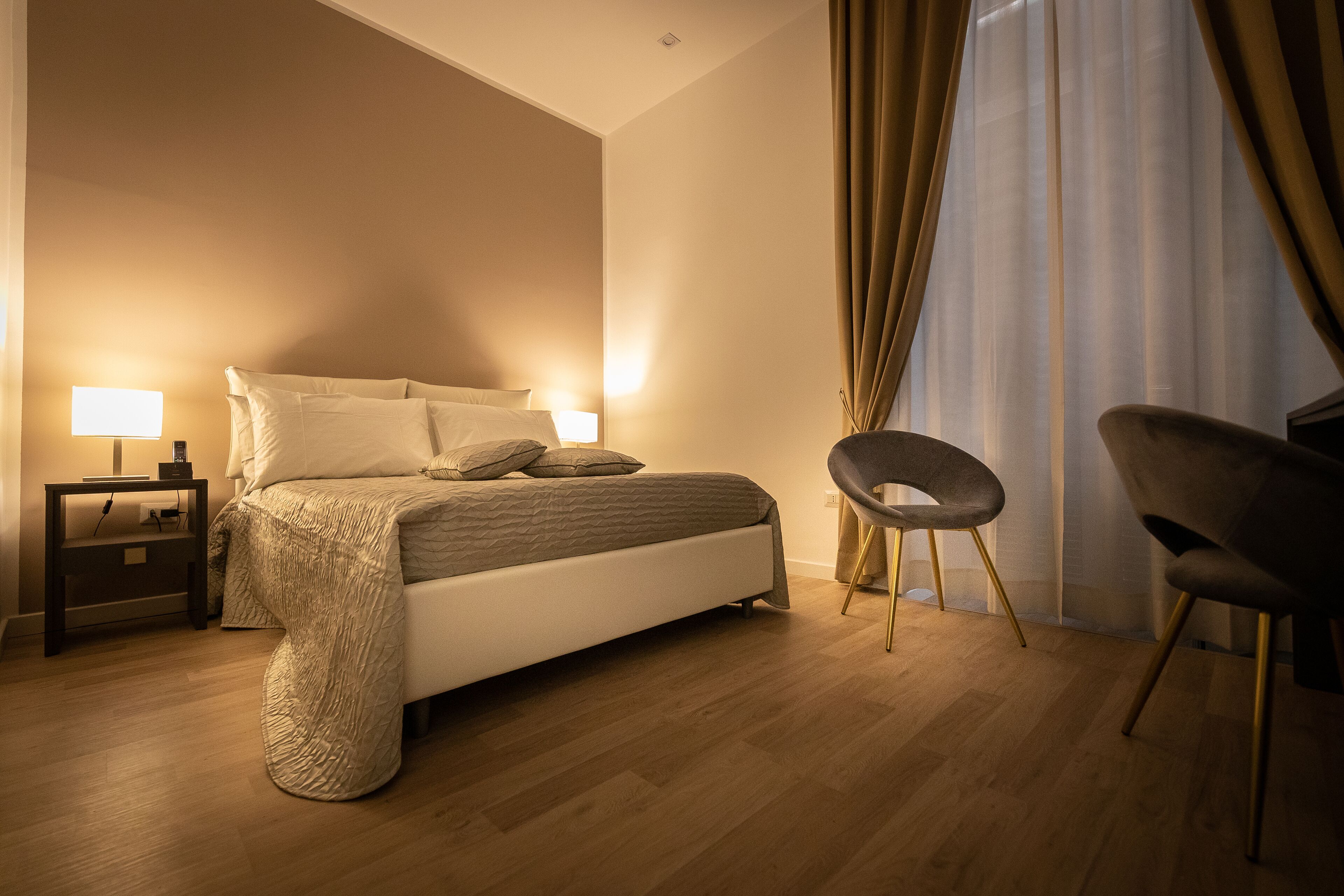 A bedroom with a bed, two bedside tables with lamps, a chair, and a large window with curtains.