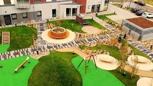Een speeltuin met groen gras, een ronde houten speelconstructie en een wipkip.