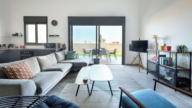 Una sala de estar moderna con un sofá gris, una mesa de centro blanca y un sillón azul. Cuenta con una zona de cocina con microondas y televisión. La habitación tiene un gran ventanal corredero que da a un patio exterior con mesa y sillas.