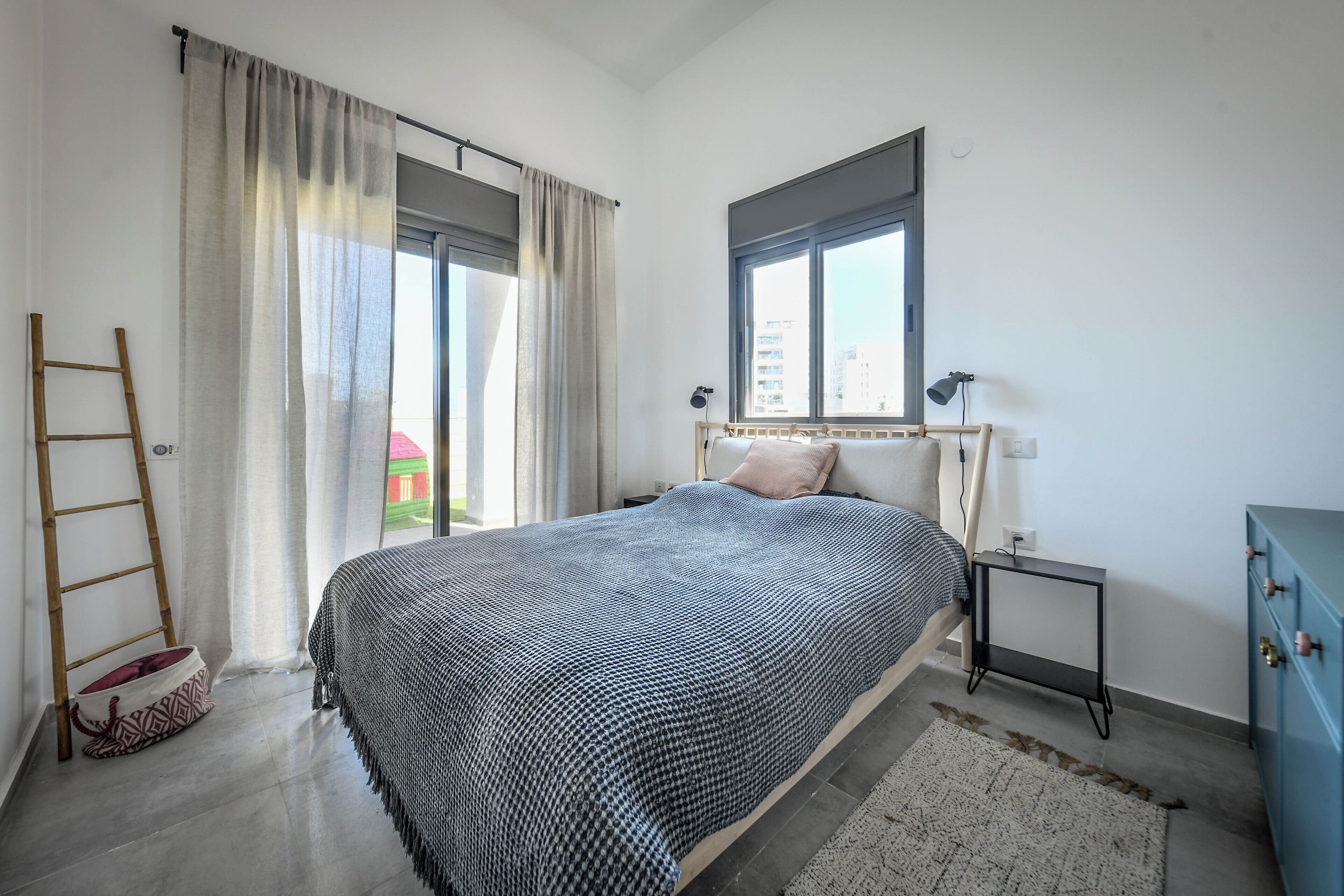 A bedroom with a bed, a nightstand, a dresser, a window, and a view of buildings outside.