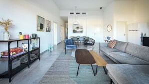 Una sala moderna con un sofá gris, una mesa de centro de madera y una consola negra con espacio de almacenamiento. En el fondo, un área de comedor con una silla azul y una mesa blanca.