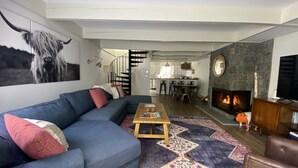 A modern living room with a blue sectional sofa, a wooden coffee table, and a patterned rug. There is a fireplace, a dining area, and a kitchen in the background.