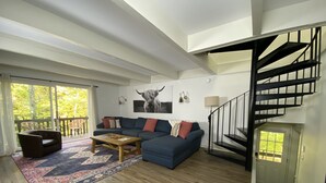 A modern living room with a blue sectional sofa, a wooden coffee table, and a patterned rug. A black staircase leads to an upper level.