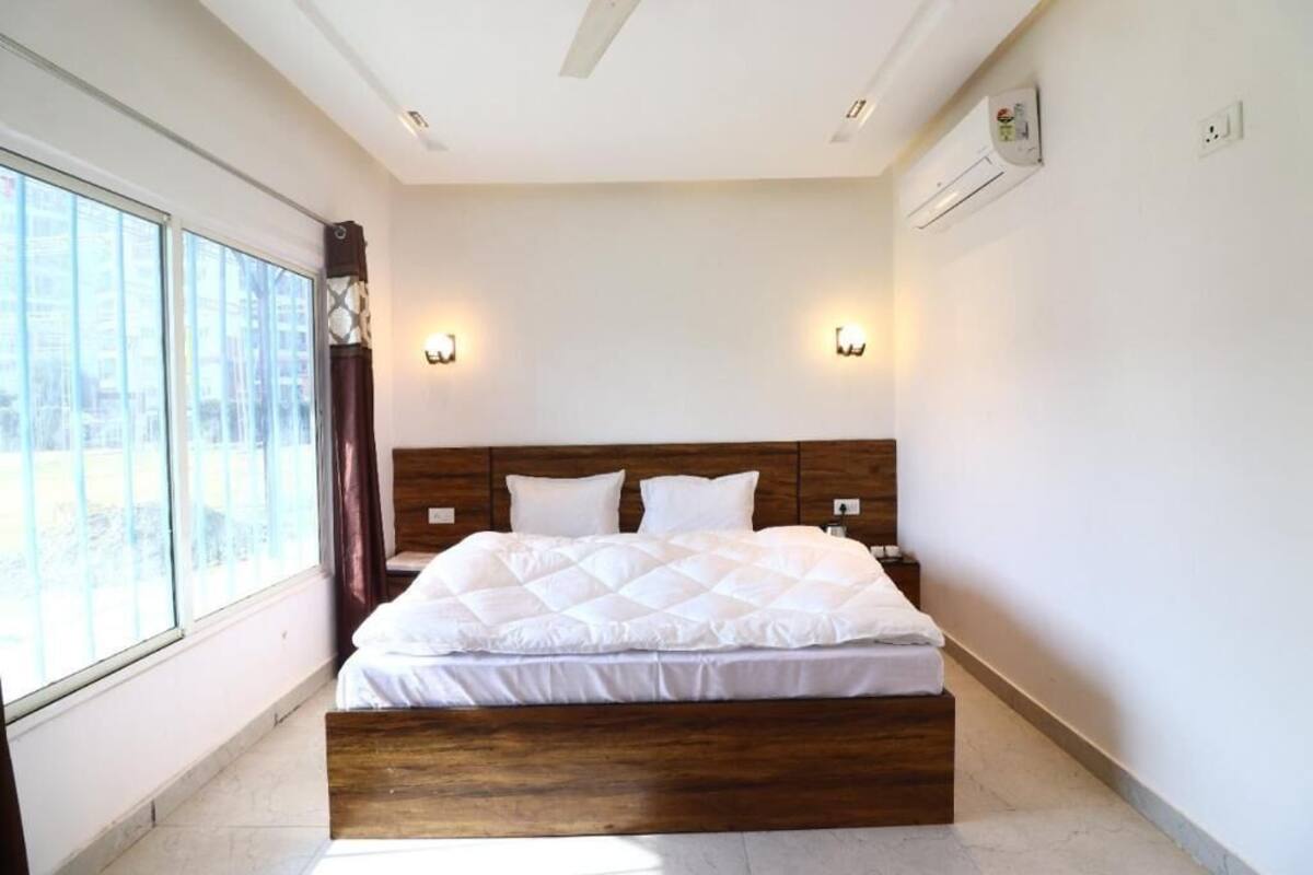 A bedroom with a large bed, two bedside tables, a window with curtains, and an air conditioning unit on the wall.