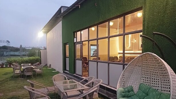 An outdoor seating area with wicker furniture and a hanging chair, a glass table, and a view of the interior through large windows. - Vrinda Resort by Dcrest  (Mathura)