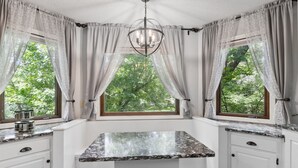 Una cocina con encimera de mármol, armarios blancos y vistas a árboles por las ventanas.