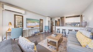 A modern living room with a sofa, coffee table, and a TV. There is a kitchen area with a fridge and bar stools.