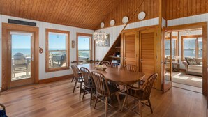 A spacious dining area with a large wooden table and chairs, a chandelier, and large windows offering ocean views. - D124- Classic Duck Oceanfront - 4 Brs Outstanding Ocean Views & Comm. Pool (Duck)