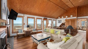 A spacious living room with a wooden ceiling, large windows, a fireplace, and a television mounted on the wall. - D124- Classic Duck Oceanfront - 4 Brs Outstanding Ocean Views & Comm. Pool (Duck)