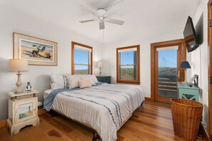 A bedroom with a bed, a ceiling fan, a framed painting, two windows, a television, and a wicker basket.
