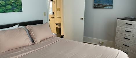 A bedroom with a bed, a dresser, a painting, and a door leading to a bathroom.