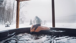 Eine Person entspannt in einer Badewanne, durch große Fenster ist eine verschneite Landschaft zu sehen.