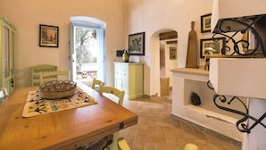 Comedor con una mesa de madera, sillas, una cesta tejida y un camino de mesa estampado. Se observa una puerta que da al exterior, un mueble con un florero y un estante con adornos.