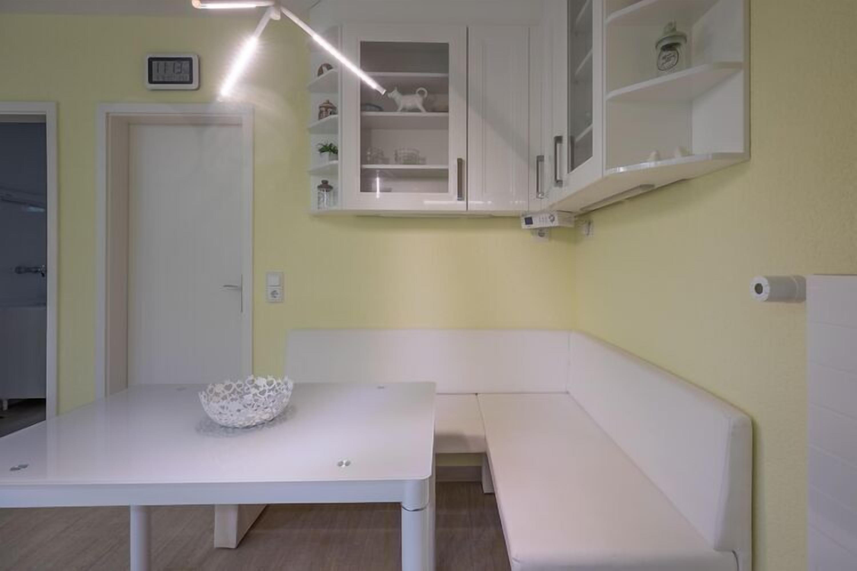 A small, bright room with a white table and bench, a cabinet with shelves, and a door.