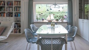 Comedor con mesa de vidrio, sillas de metal, una estantería y vista a un jardín a través de un ventanal.