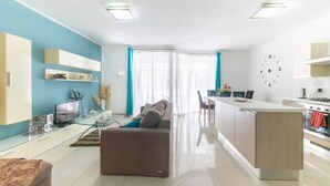 A modern living room with a sofa, TV, and a glass coffee table. There is a kitchen area with a stove and a dining table. - ☆Amazing Apart.w/side Sea View☆8 (San Pawl il-Bahar)