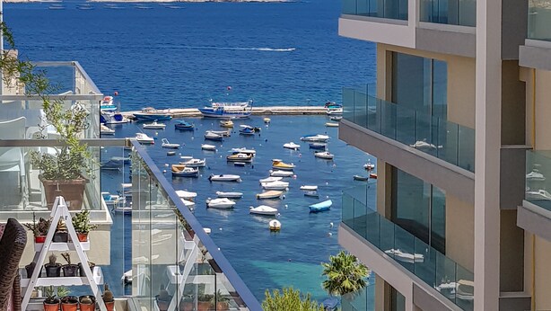 A balcony with a view of the sea, boats, and a modern building with glass railings. - ☆Amazing Apart.w/side Sea View☆8 (San Pawl il-Bahar)