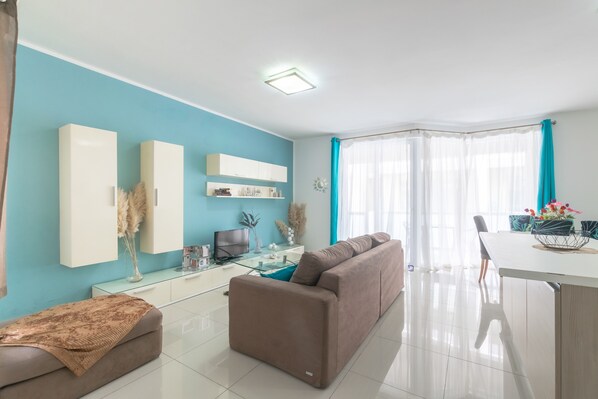 A modern living room with a sofa, a glass coffee table, and a TV. There is a dining area with a table and chairs. - ☆Amazing Apart.w/side Sea View☆8 (San Pawl il-Bahar)
