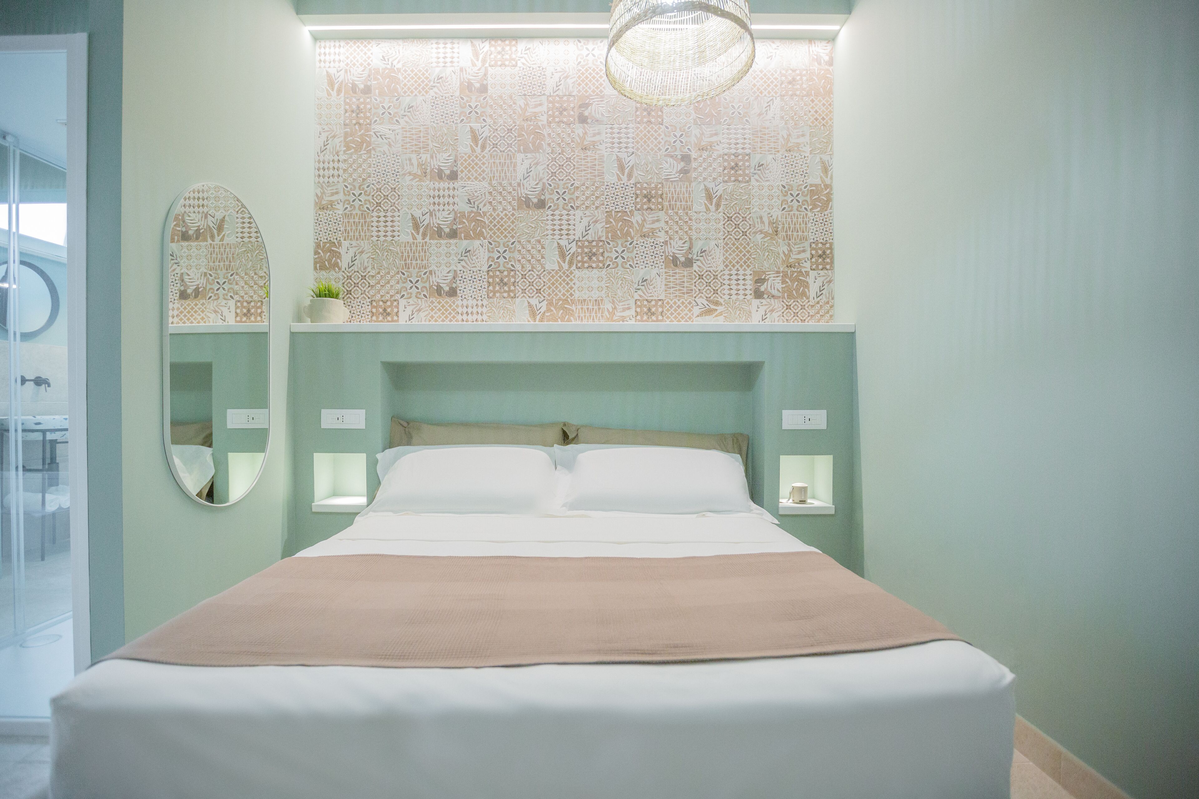 A neatly made bed with a beige blanket, a large mirror, and a patterned wall above it.
