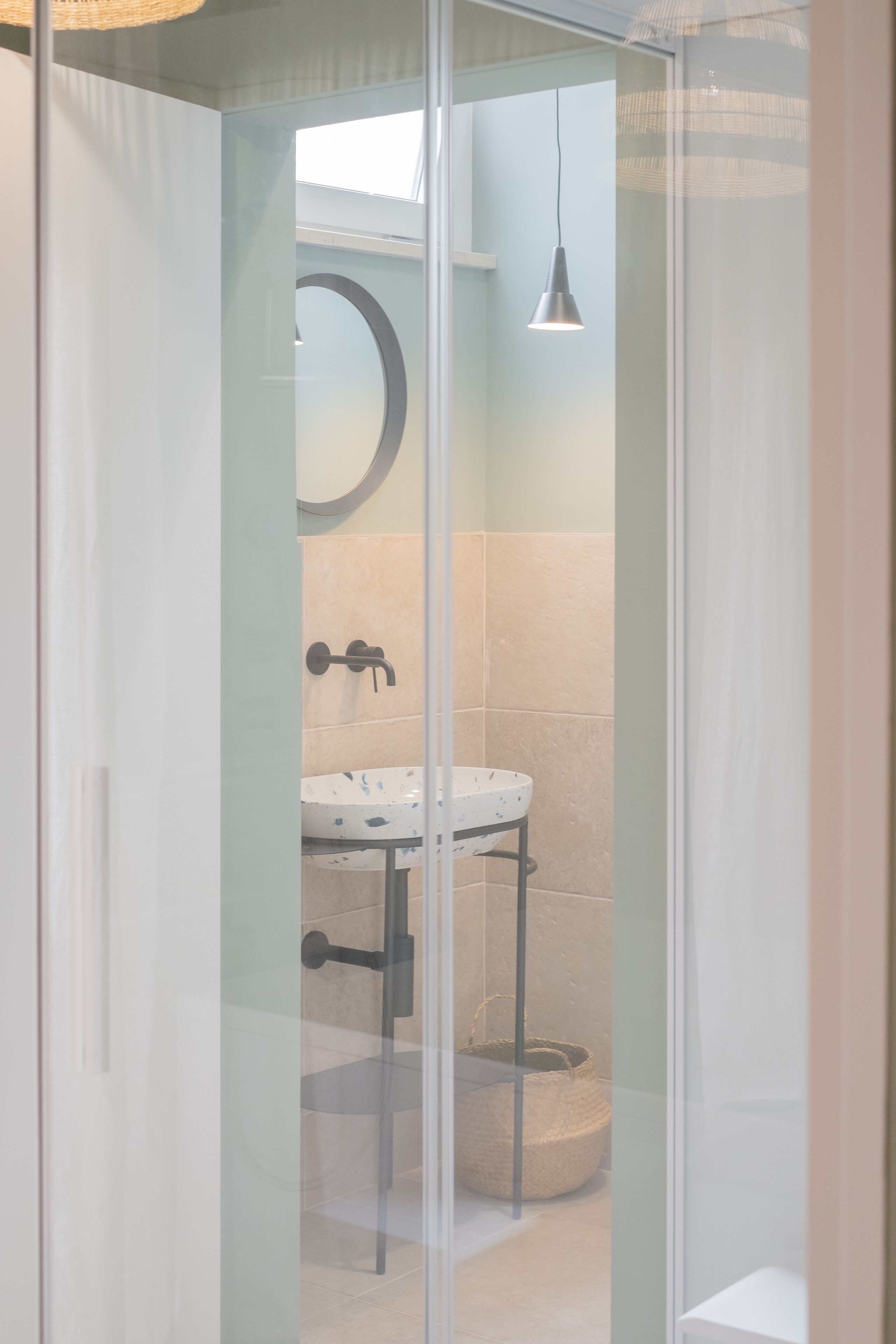 Un espace douche clos par des parois de verre, avec un lavabo blanc, un miroir rond et un luminaire suspendu.