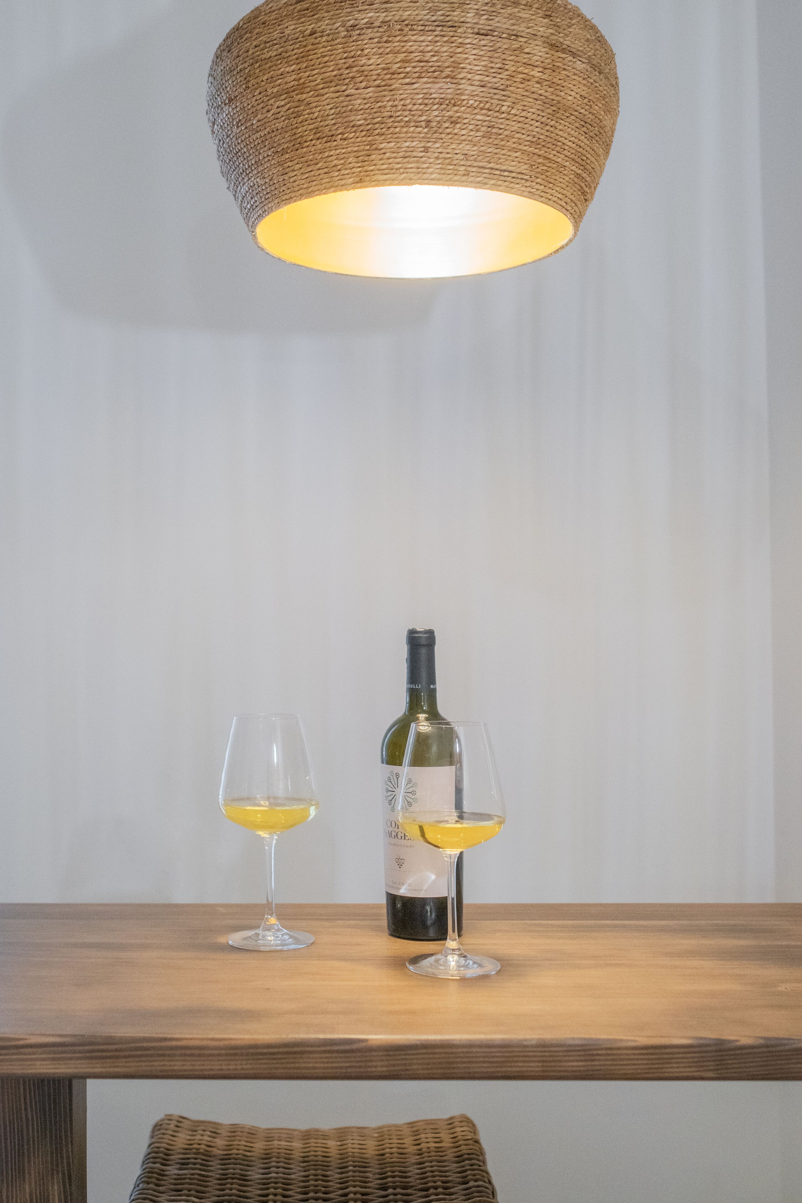 Una mesa con dos copas de vino y una botella, bajo una lámpara de fibra trenzada.